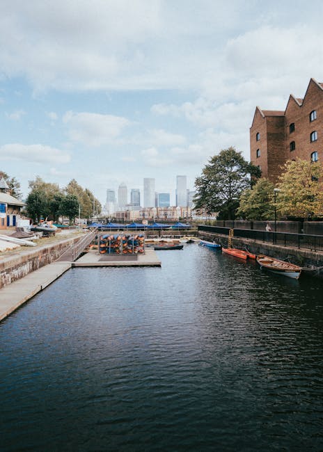 A panoramic view of the London skyline near Canary Wharf in the Docklands, featuring several modern high-rise office buildings and residential flats. In the foreground, there is a waterfront with a ca